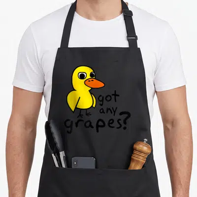 Song duck apron - Yellow cartoon duck with orange beak next to stacked got any grapes text in black font — shown on a black cotton canvas apron against a white background