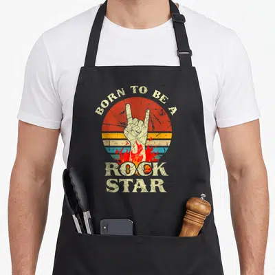 Rock star apron - Distressed hand making rock sign in front of striped sun with flames and text — shown on a black cotton canvas apron against a white background