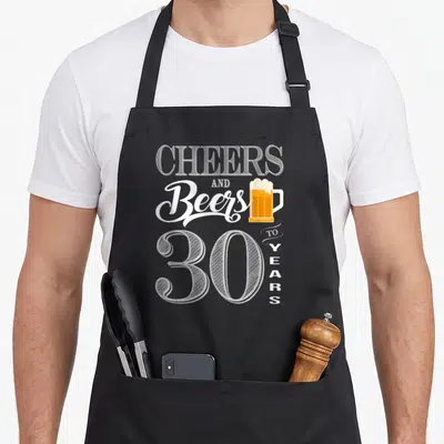 30th cheer birthday apron - Stacked CHEERS AND Beers 30 YEARS text with beer mug in white yellow orange — shown on a black cotton canvas apron against a white background