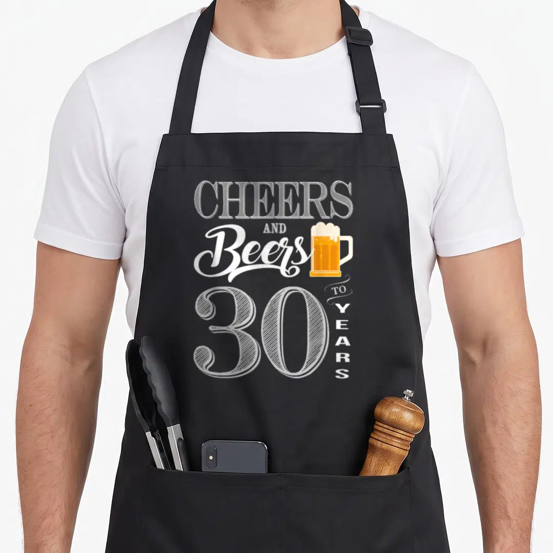 30th cheer birthday apron - Stacked CHEERS AND Beers 30 YEARS text with beer mug in white yellow orange — shown on a black cotton canvas apron against a white background