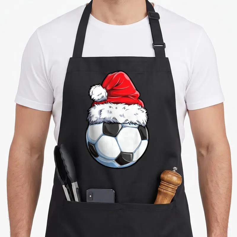 Soccer santa aprons - Cartoon soccer ball wearing a red and white Santa hat in black, white, and red art style — shown on a black cotton canvas apron against a white background