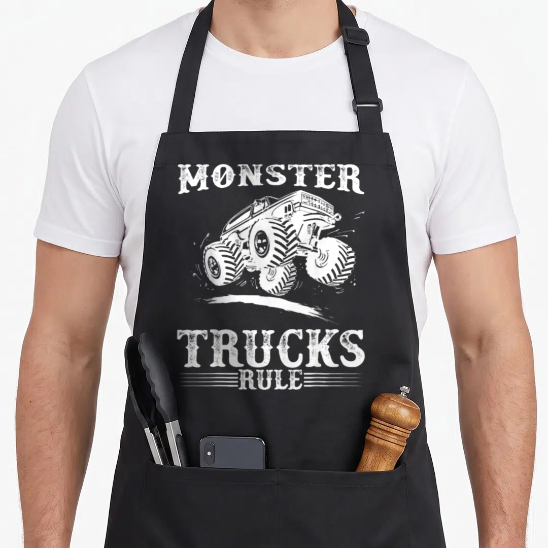 Jumping truck apron - White cartoon monster truck centered between MONSTER and TRUCKS RULE text — shown on a black cotton canvas apron against a white background
