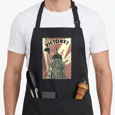 Propoganda collectible apron - Dalek centered in red frame with cream rays and TO, VICTORY! text in distressed style — shown on a black cotton canvas apron against a white background