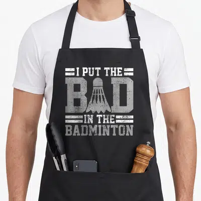 Player badminton quotes apron - White distressed text I PUT THE BAD IN THE BADMINTON with shuttlecock and stripes — shown on a black cotton canvas apron against a white background