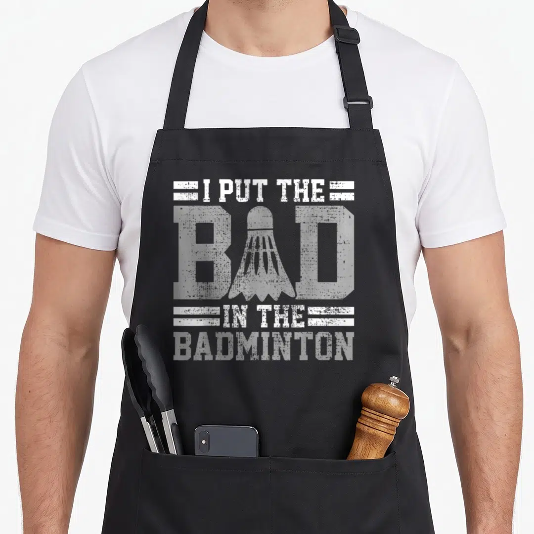Player badminton quotes apron - White distressed text I PUT THE BAD IN THE BADMINTON with shuttlecock and stripes — shown on a black cotton canvas apron against a white background