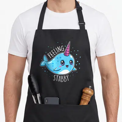 Feeling kawaii apron - Blue cartoon narwhal with pink horn and white text reading FEELING STABBY surrounded by dots — shown on a black cotton canvas apron against a white background