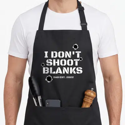 Baby blank apron - White stenciled text I Don't Shoot Blanks Dad Est 2022 with four black bullet holes — shown on a black cotton canvas apron against a white background