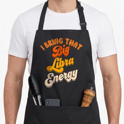 Energy birthday apron - I Bring That Big Libra Energy text in orange yellow and peach retro script — shown on a black cotton canvas apron against a white background