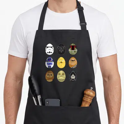 Original easter egg apron - Nine cartoon egg portraits of characters in white, black, yellow, brown, green, and blue — shown on a black cotton canvas apron against a white background