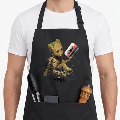 Guardian galaxies apron - Baby Groot holding Awesome Mix Vol 2 cassette with unspooled black tape swirling around — shown on a black cotton canvas apron against a white background