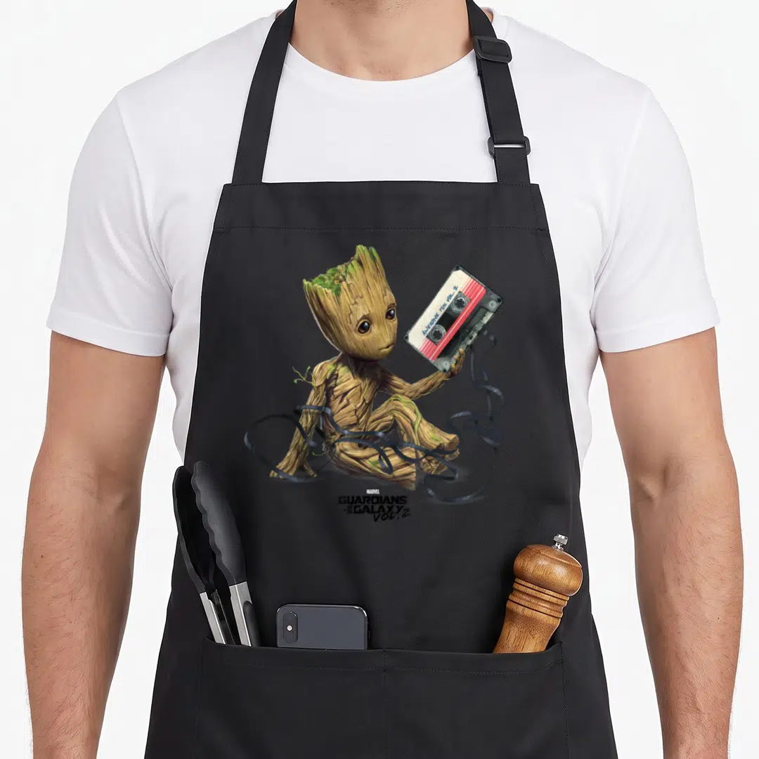 Guardian galaxies apron - Baby Groot holding Awesome Mix Vol 2 cassette with unspooled black tape swirling around — shown on a black cotton canvas apron against a white background