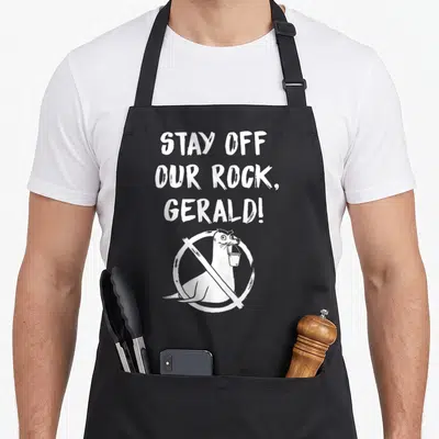 Finding stay apron - White text STAY OFF OUR ROCK GERALD above seal with bucket in prohibition sign — shown on a black cotton canvas apron against a white background