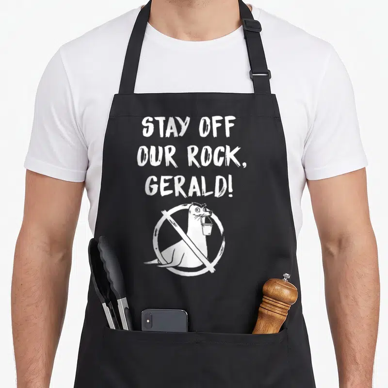 Finding stay apron - White text STAY OFF OUR ROCK GERALD above seal with bucket in prohibition sign — shown on a black cotton canvas apron against a white background