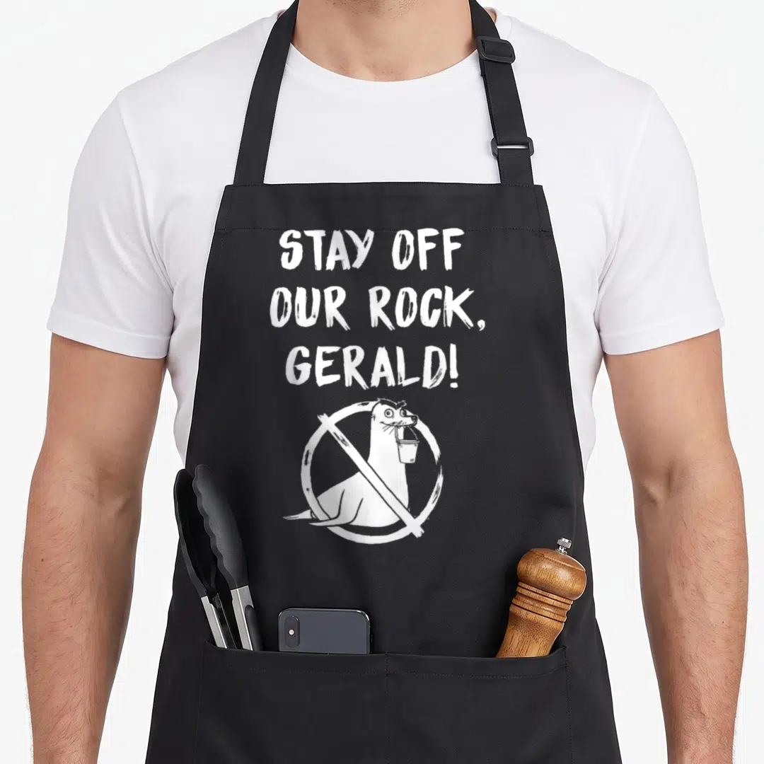 Finding stay apron - White text STAY OFF OUR ROCK GERALD above seal with bucket in prohibition sign — shown on a black cotton canvas apron against a white background