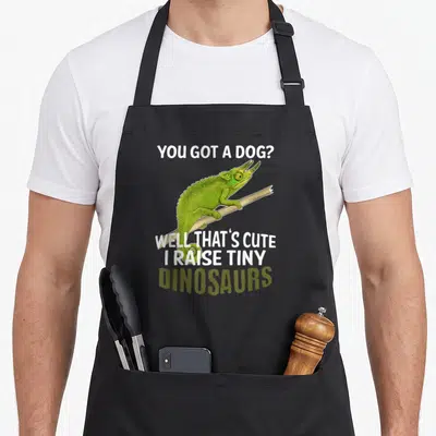 Jackson pet apron - Green chameleon on branch with white and olive text about raising dinosaurs — shown on a black cotton canvas apron against a white background