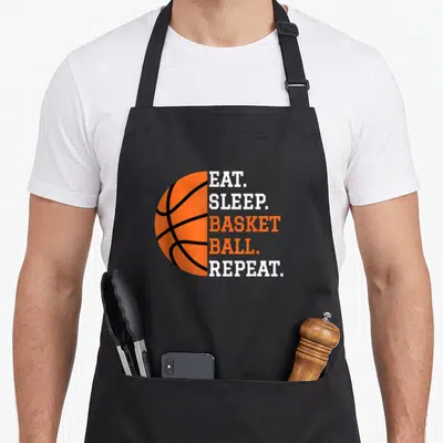 Eat basketball player apron - Half orange basketball with stacked EAT SLEEP BASKET BALL REPEAT text — shown on a black cotton canvas apron against a white background