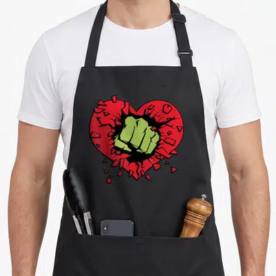 Smash couple apron - Green fist punching through a shattered red heart in cartoon style — shown on a black cotton canvas apron against a white background