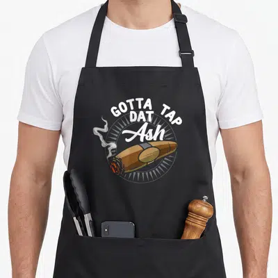 Tap my therapy apron - Gotta Tap Dat Ash text over brown cigar with grey smoke on circular background — shown on a black cotton canvas apron against a white background