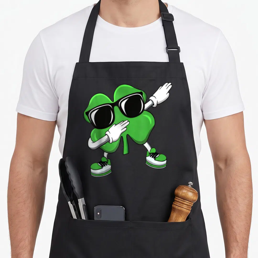 Shamrock dab st patrick day aprons - Green cartoon shamrock wearing sunglasses and sneakers striking a pose — shown on a black cotton canvas apron against a white background