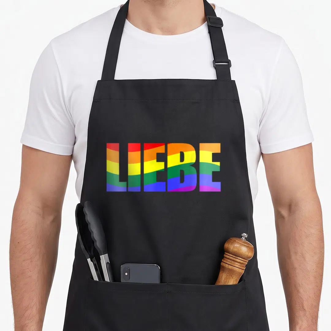 Liebe meme apron - LIEBE text filled with red orange yellow green blue purple rainbow stripes — shown on a black cotton canvas apron against a white background