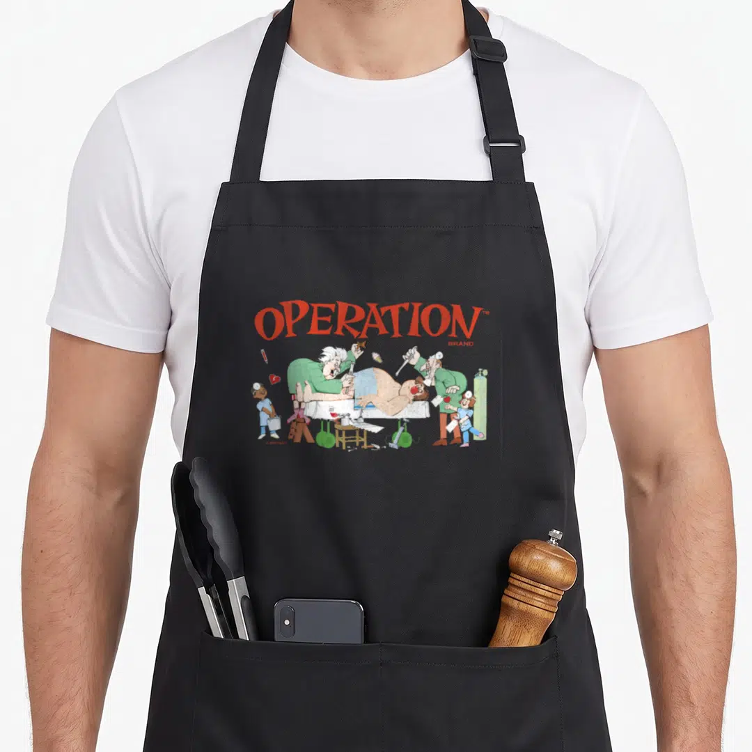 Operation surgeon apron - Cartoon surgeons operating on a patient with red arched text reading OPERATION — shown on a black cotton canvas apron against a white background