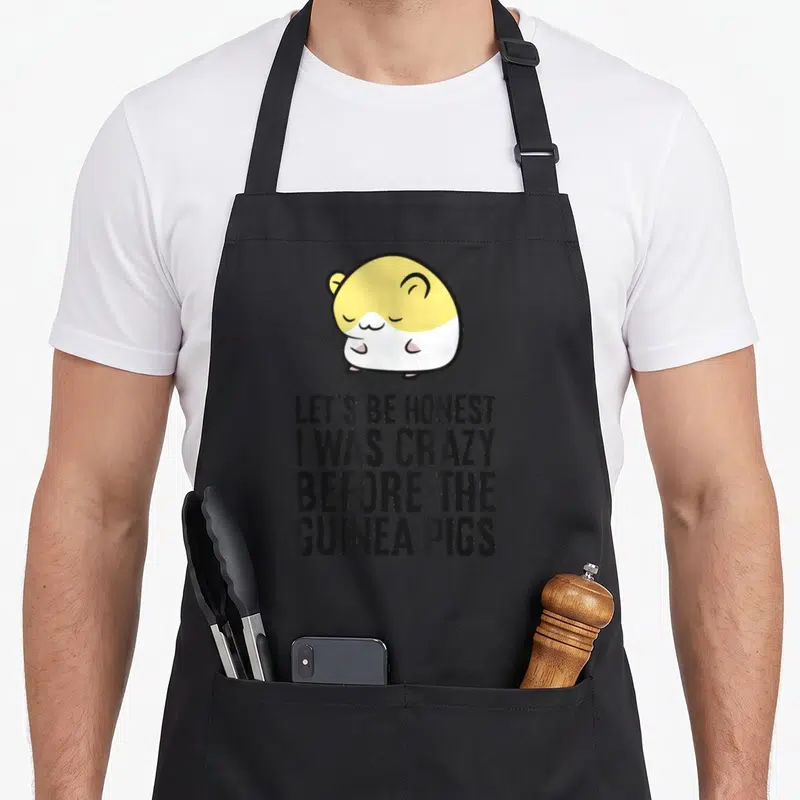 Honest crazy guinea man apron - Cartoon guinea pig in yellow and white above four lines of stacked black text — shown on a black cotton canvas apron against a white background