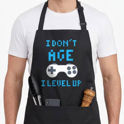 Funny gaming apron - Light blue pixel text I Don't Age I Level Up surrounding a white game controller on a grey — shown on a black cotton canvas apron against a white background