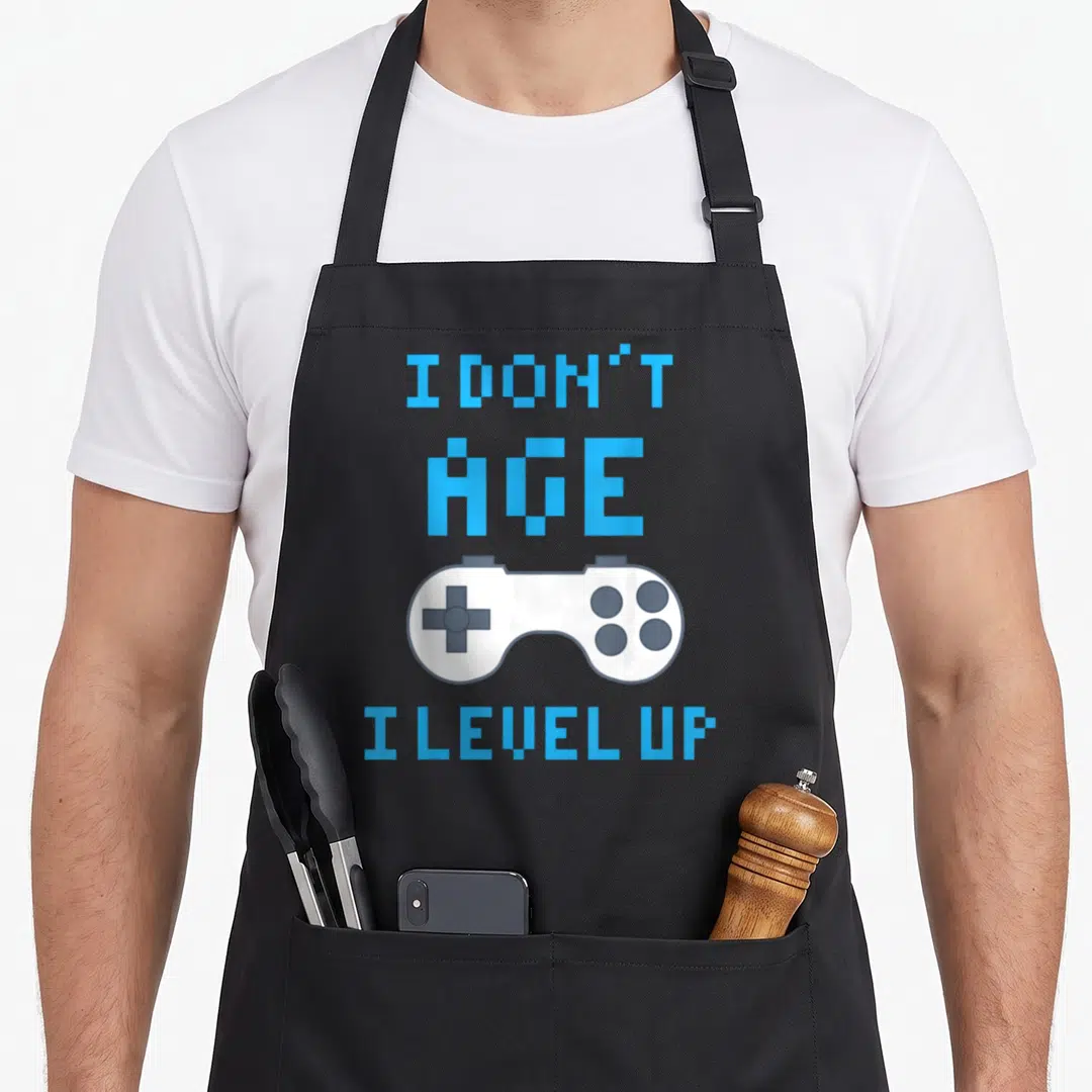 Funny gaming apron - Light blue pixel text I Don't Age I Level Up surrounding a white game controller on a grey — shown on a black cotton canvas apron against a white background