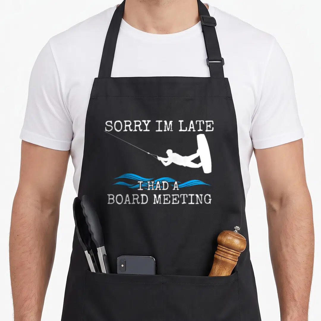 Wakeboard wakeboarding apron - White kitesurfer silhouette with blue waves and sorry im late board meeting text — shown on a black cotton canvas apron against a white background