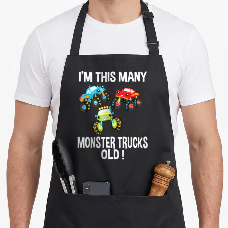 Birthday many truck apron - Three cartoon monster trucks in blue red and green with text I'M THIS MANY MONSTER TRUCKS — shown on a black cotton canvas apron against a white background