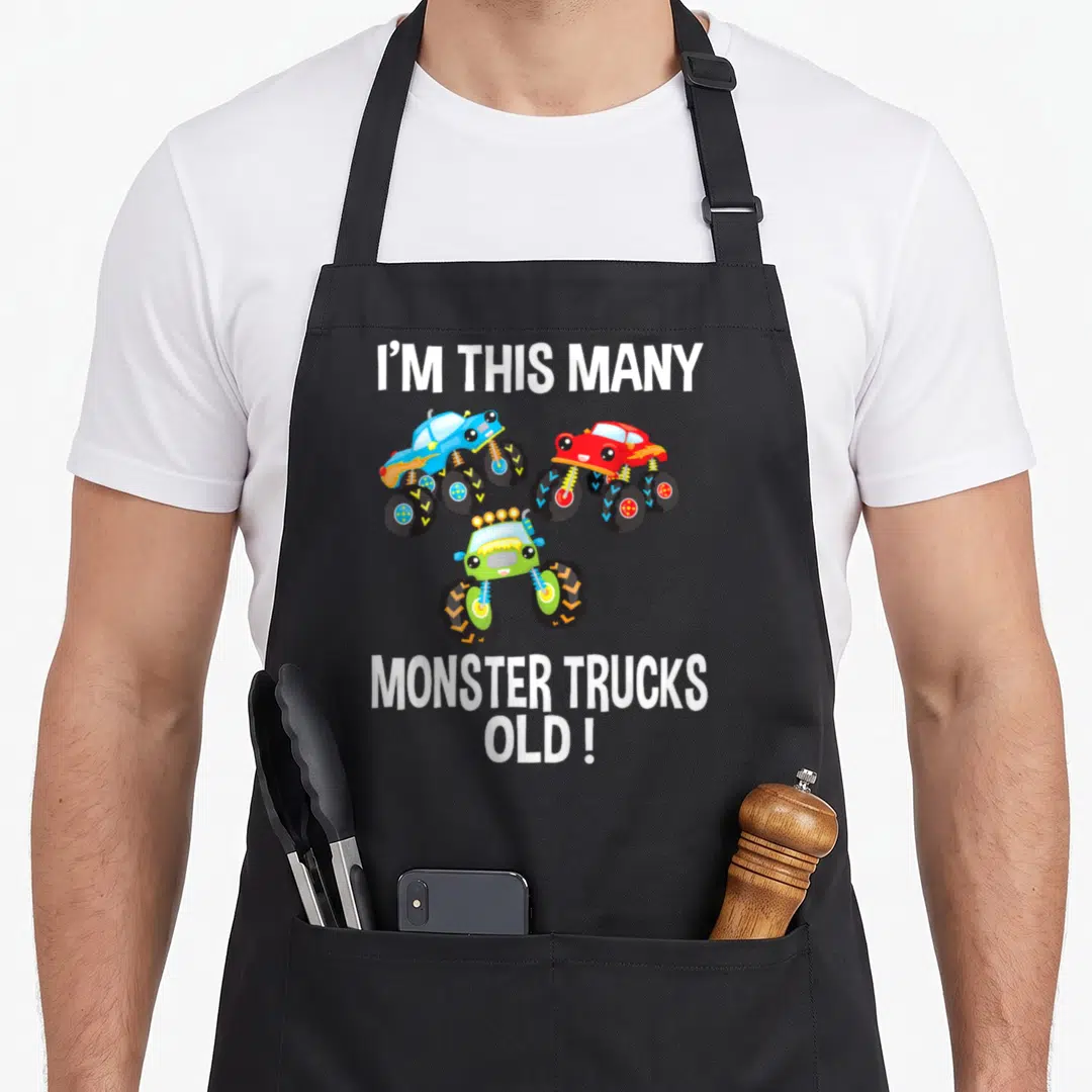 Birthday many truck apron - Three cartoon monster trucks in blue red and green with text I'M THIS MANY MONSTER TRUCKS — shown on a black cotton canvas apron against a white background