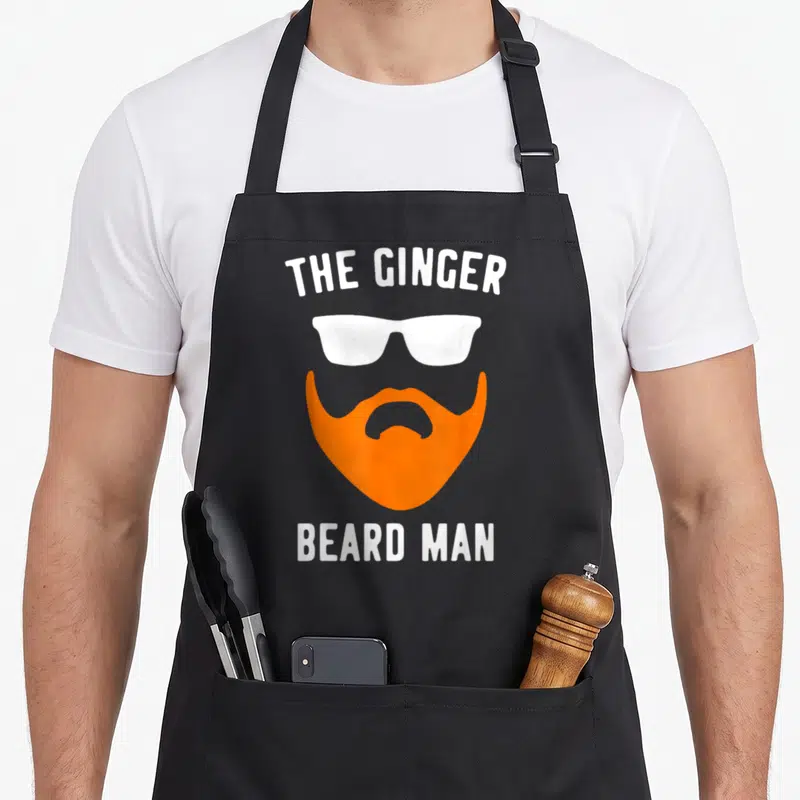 Ginger beard apron - White sunglasses above orange beard with THE GINGER BEARD MAN text — shown on a black cotton canvas apron against a white background
