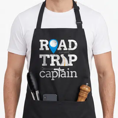 Captain road apron - Distressed white text ROAD TRIP captain with blue map pin, orange road, and airplane graphics — shown on a black cotton canvas apron against a white background