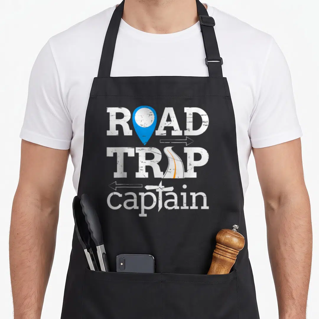 Captain road apron - Distressed white text ROAD TRIP captain with blue map pin, orange road, and airplane graphics — shown on a black cotton canvas apron against a white background