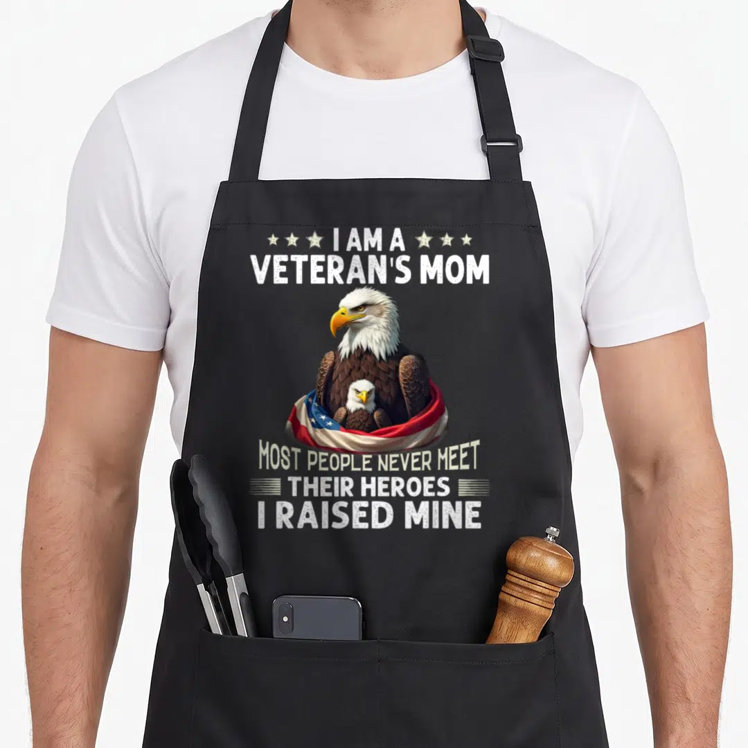 Raised eagles apron - Bald eagle holding chick with flag and veteran mom text — shown on a black cotton canvas apron against a white background