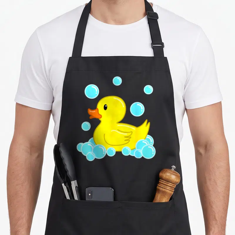 Duckie duck apron - Yellow cartoon rubber duck with orange beak and light blue soap bubbles — shown on a black cotton canvas apron against a white background