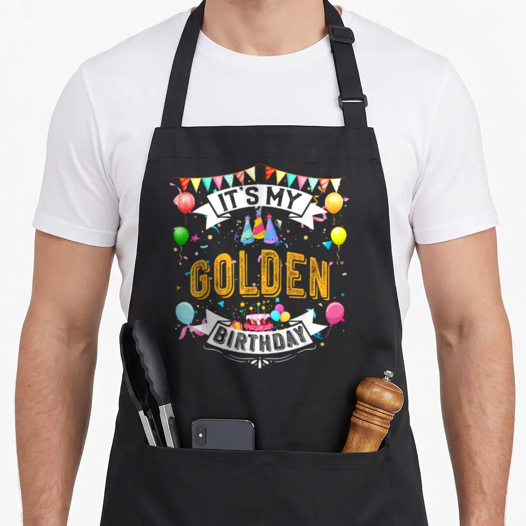 Birthday golden apron - Cartoon It's My Golden Birthday text with yellow letters, party hats, balloons, and bunting — shown on a black cotton canvas apron against a white background