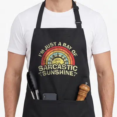 Ray sarcastic apron - Retro rainbow with smiling sun and palm trees surrounding sarcastic text — shown on a black cotton canvas apron against a white background