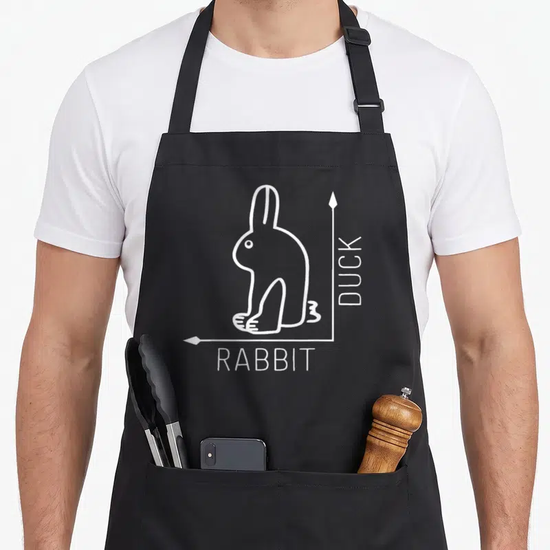 Duck rabbit apron - White line-art rabbit-duck illusion with DUCK and RABBIT text labels on black fabric — shown on a black cotton canvas apron against a white background
