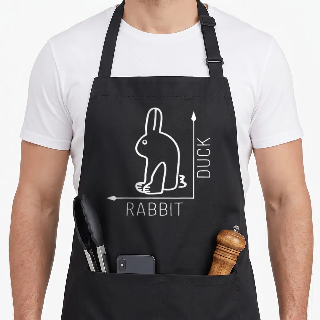 Duck rabbit apron - White line-art rabbit-duck illusion with DUCK and RABBIT text labels on black fabric — shown on a black cotton canvas apron against a white background
