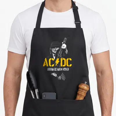 Dark wellness shot apron - AC/DC yellow logo and SHOT IN THE DARK white text over grey photo of man with guitar — shown on a black cotton canvas apron against a white background