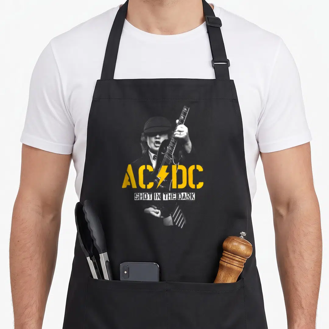 Dark wellness shot apron - AC/DC yellow logo and SHOT IN THE DARK white text over grey photo of man with guitar — shown on a black cotton canvas apron against a white background