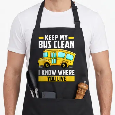 Traffic bus driver apron - Cartoon yellow school bus with text KEEP MY BUS CLEAN I KNOW WHERE YOU LIVE — shown on a black cotton canvas apron against a white background