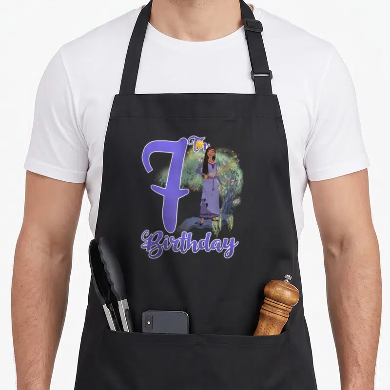 Asha birthday apron - Purple number 7, girl standing by tree, Th Birthday text in cartoon style — shown on a black cotton canvas apron against a white background