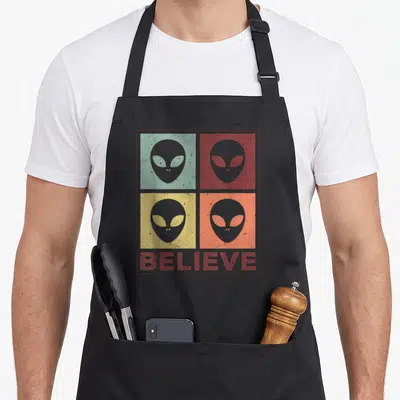 Believe believer apron - Retro alien heads in red, green, yellow, and orange squares above BELIEVE text — shown on a black cotton canvas apron against a white background
