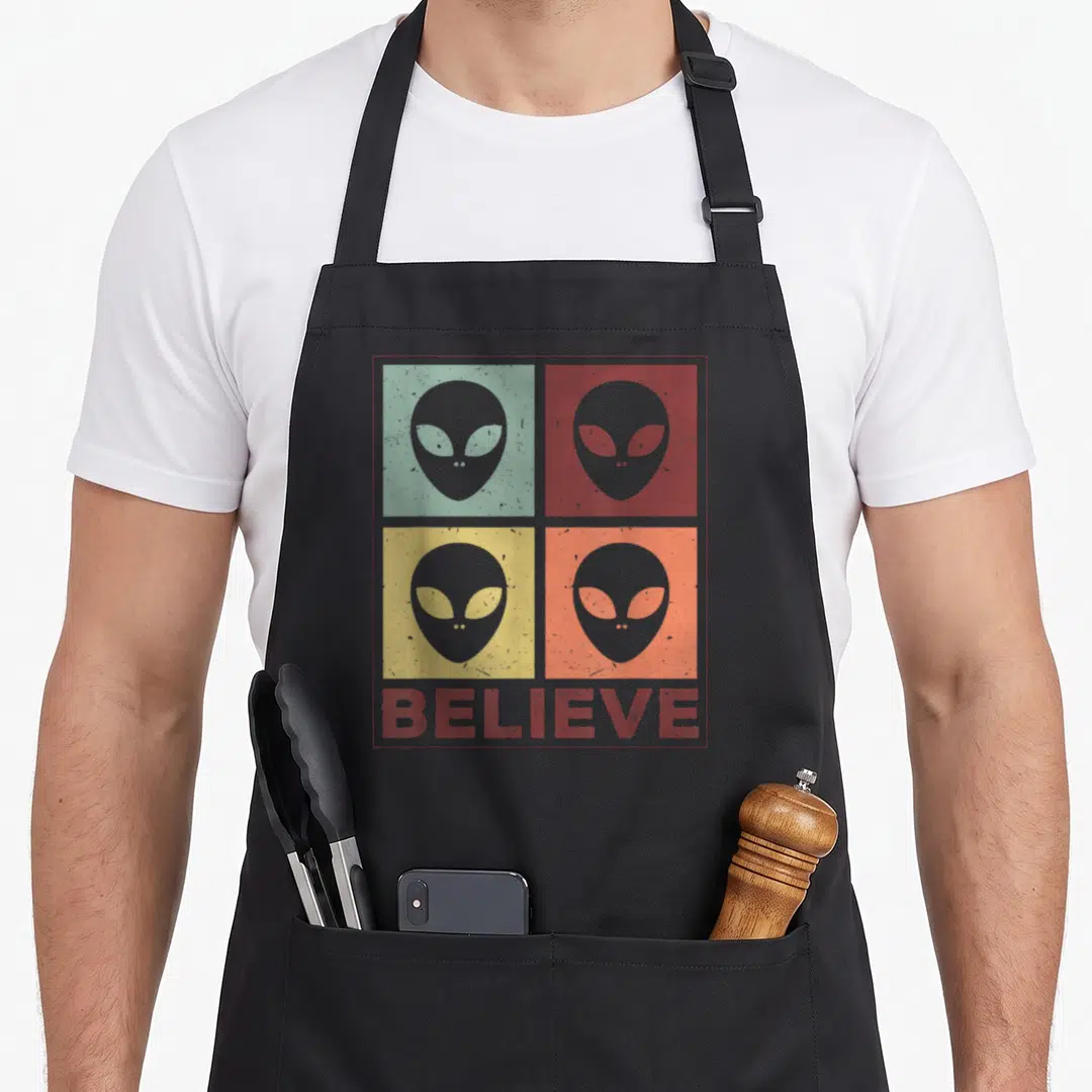 Believe believer apron - Retro alien heads in red, green, yellow, and orange squares above BELIEVE text — shown on a black cotton canvas apron against a white background