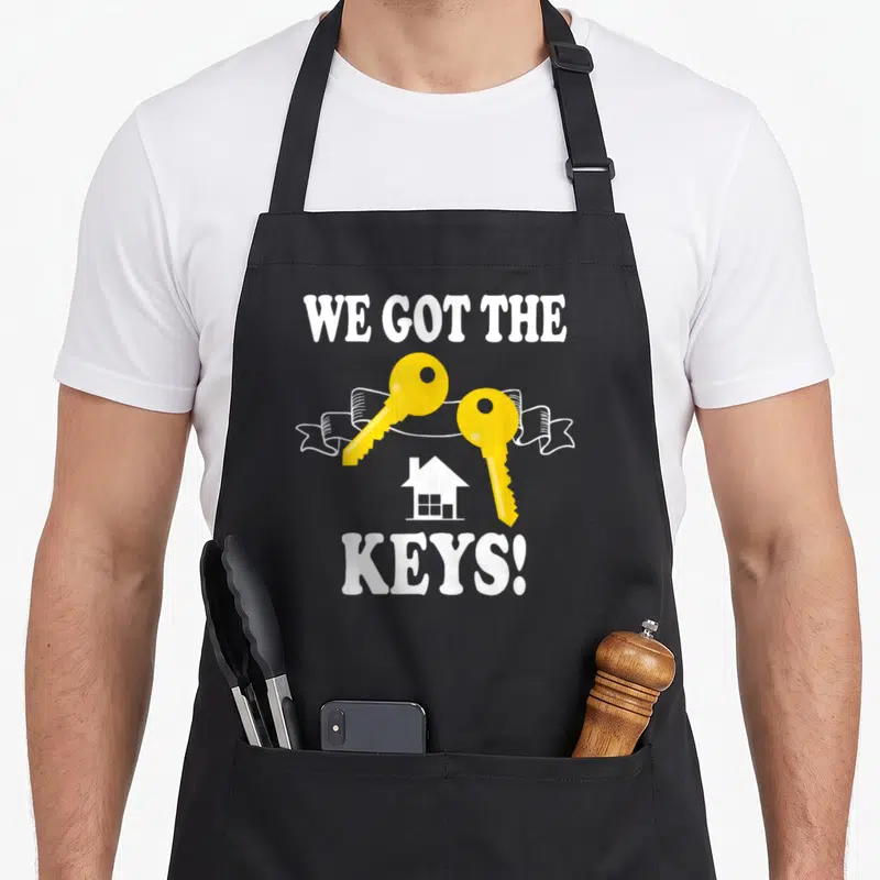 Key homeowner apron - WE GOT THE KEYS text with two yellow keys, white ribbon, and house silhouette — shown on a black cotton canvas apron against a white background