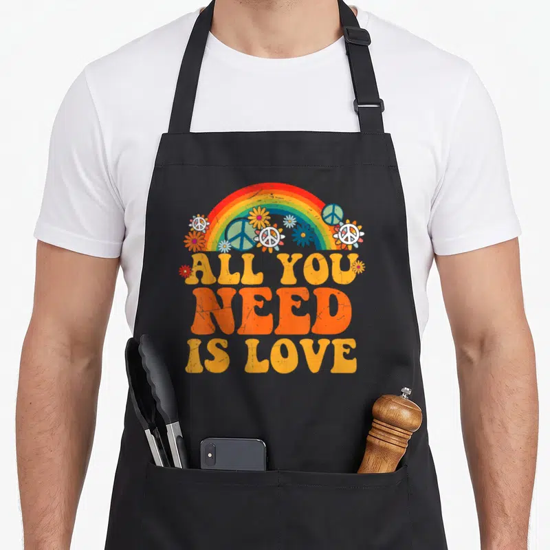 Dye hippie apron - Retro rainbow arching over flowers and peace symbols with stacked orange text reading ALL YOU NEED — shown on a black cotton canvas apron against a white background