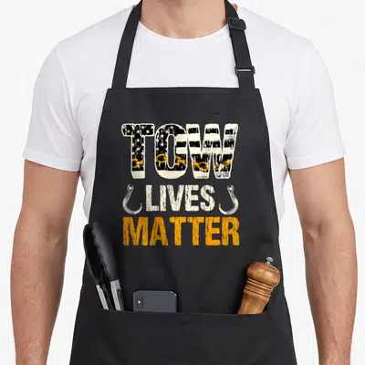 Cute tow truck apron - TOW LIVES MATTER text with chain and hooks in white orange black distressed style — shown on a black cotton canvas apron against a white background