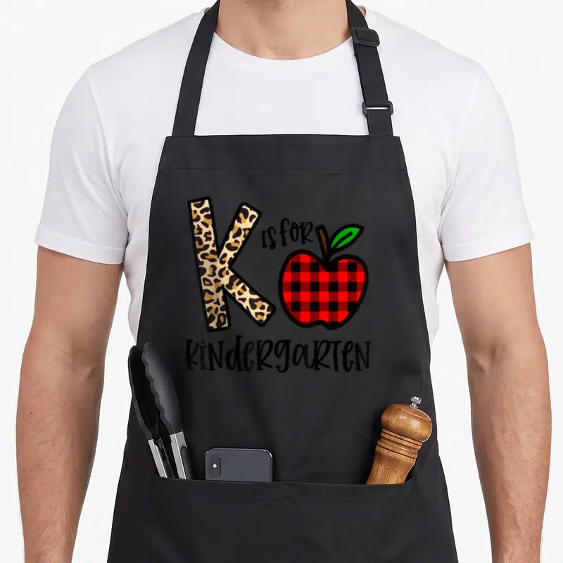 Leopard kindergarten teacher apron - Leopard print letter K next to red plaid apple and black KINDERGARTEN text — shown on a black cotton canvas apron against a white background